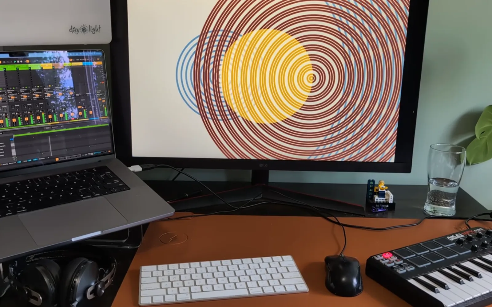 An audio visualizer on a monitor at a desk: concentric orange, dark red and blue rings radiating from a solid yellow disc on a cream background, with a laptop running a digital audio workstation and a MIDI keyboard alongside.